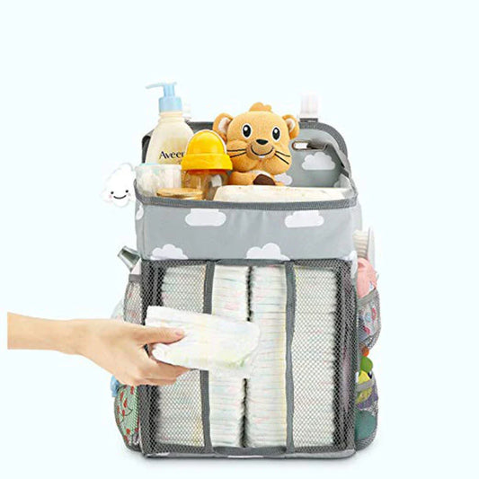 A gray crib-side organizer hanging on a white crib, storing various baby items like diapers, bottles, and toys.