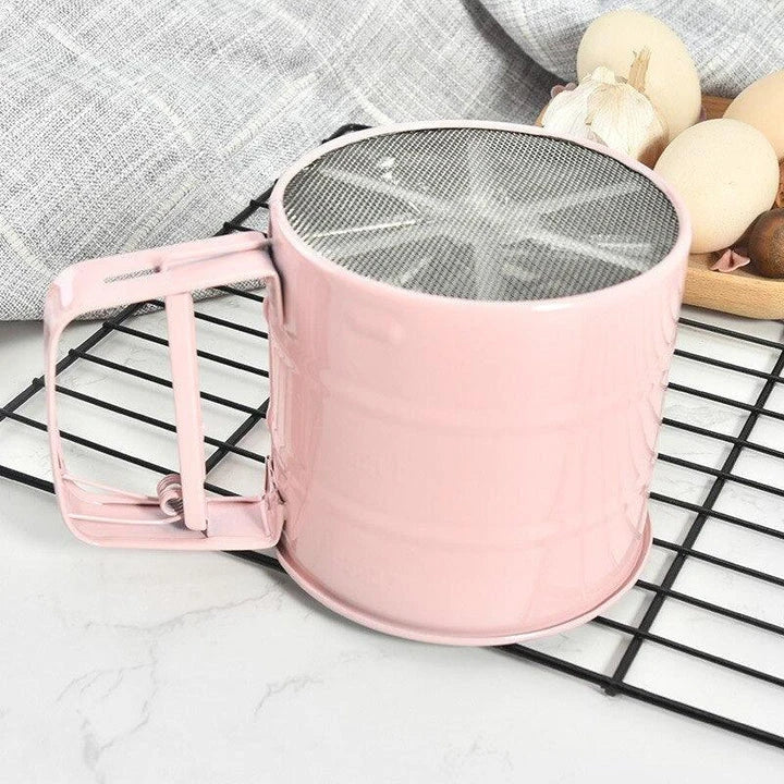 Stainless steel handheld baking flour sifter in a soft pink colour, designed for efficient sifting and aeration of dry ingredients for light and fluffy baked goods.