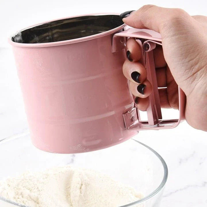 Stainless steel handheld baking flour sifter in a soft pink colour, designed for efficient sifting and aeration of dry ingredients for light and fluffy baked goods.