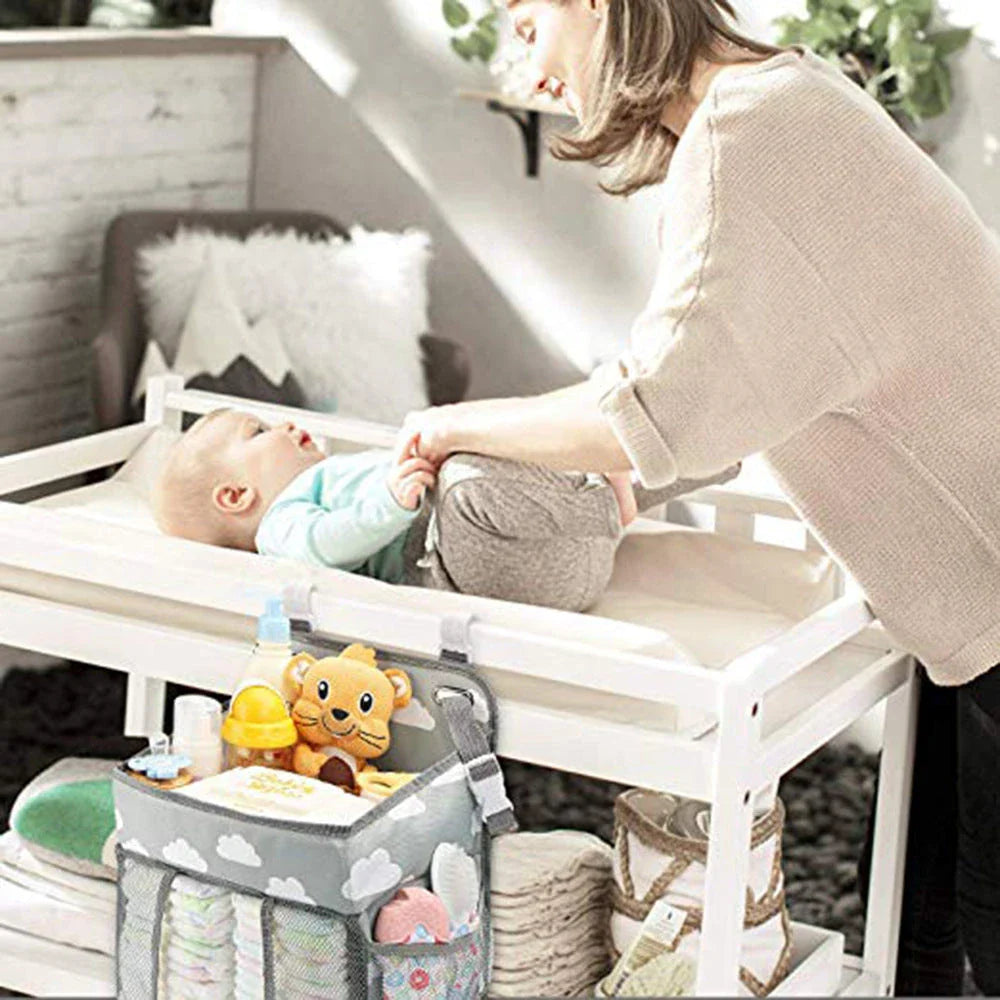 A gray crib-side organizer hanging on a white crib, storing various baby items like diapers, bottles, and toys.